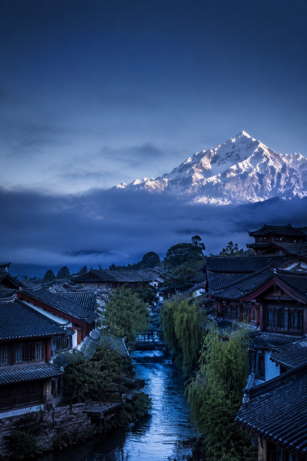 雪山云雾下，丽江木构屋顶与水道向远处延展的意象画面